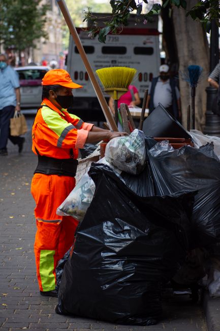 A street scene featuring a sanitation worker wearing a beige cap, blue uniform, face mask, and gloves, walking along the pavement while pushing a wheeled cart loaded with a large orange trash bin and 